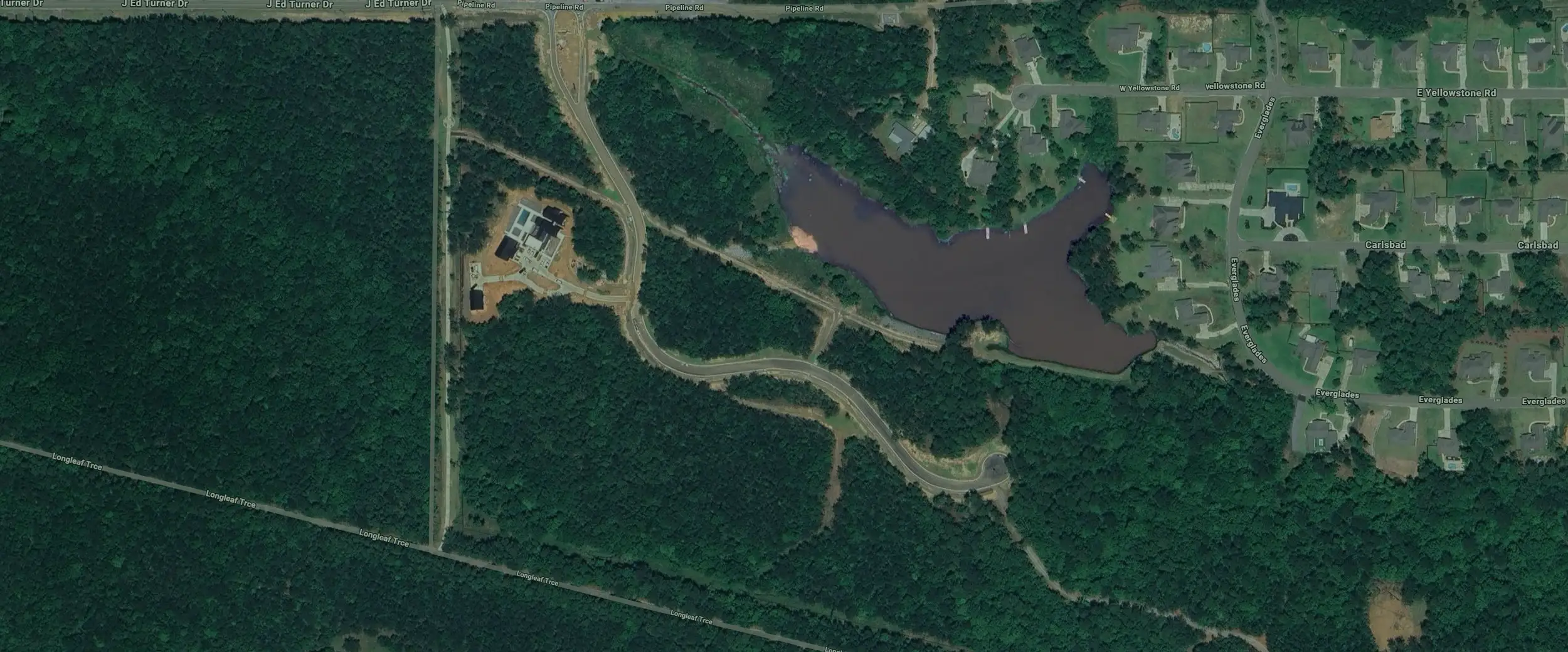 Aerial view of a wooded area with winding roads, a small body of water near houses, and a construction site surrounded by dense forest. Residential neighborhood is visible in the upper right.